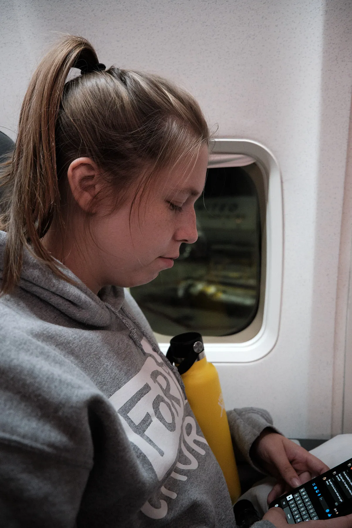 Johnna sitting in the window seat, she has a water bottle in her arms and she is looking down at her phone sending messages as we wait for the plane to leave the gate.